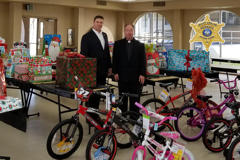 An Officer and a Priest standing among donations, wrapped gifts and bicycles.